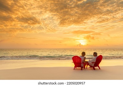 Senior Caucasian American Couple Enjoying Relaxing Beach Vacation Sitting On Red Chairs With Ocean View At Sunset Bahamas