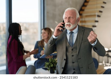 A senior Caucasian businessman in professional attire actively communicates on a phone call while gesturing, as diverse colleagues engage in the background of a modern office. - Powered by Shutterstock