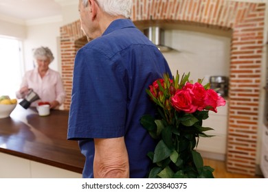 Senior Caucasian Couple Spending Time At Home Together, The Man Holding A Bouquet Of Flowers Behind His Back, Before Giving Them To His Wife. Isolating During Coronavirus Covid 19 Quarantine Lockdown.