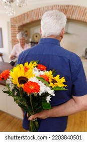 Senior Caucasian Couple Spending Time At Home Together, The Man Holding A Bouquet Of Flowers Behind His Back, Before Giving Them To His Wife. Isolating During Coronavirus Covid 19 Quarantine Lockdown.