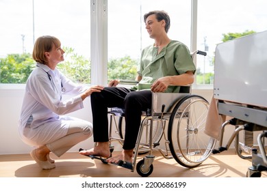 Senior Caucasian Doctor Woman Sit And Talk To Young Patient Man Who Sit On Wheelchair In Room With Glass Window And Warm Light. Doctor Try To Pacify The Patient Who Affect From Pain Of The Disease.