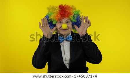Senior caucasian female clown in colorful wig and blue bow tie making silly faces, fool around. Elderly woman having fun, smiling, looking at camera. Copy space. Halloween. Yellow background