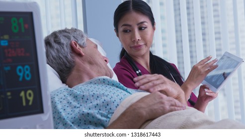 Senior Caucasian Male Patient In Hospital Bed Asking Questions About Brain Scans To Japanese Woman Doctor. Medical Professional Showing Medical Images To Older Man On Tablet Screen