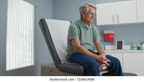 Senior Caucasian Male Patient Waiting Patiently For Doctor While Sitting On Exam Room Table. Older Man Going To Regular Appointment For Annual Check Up