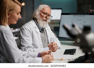 Senior Caucasian man with beard and glasses discussing research data with young adult Caucasian woman in scientific laboratory, computer monitor displaying scientific graphs