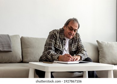A Senior Caucasian Man In His 70s Sitting At A Table Or Desk, Writing On A Notepad At Home. Senior Grey Hair Man Sitting In The Living Room And Writing In His Diary. Older Man Taking Notes At Home. 