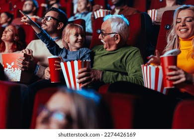 A senior Caucasian man is with his little granddaughter watching a movie in the cinema. They are bonding over the experience with popcorn as she's pointing to the big screen to him. - Shutterstock ID 2274426169