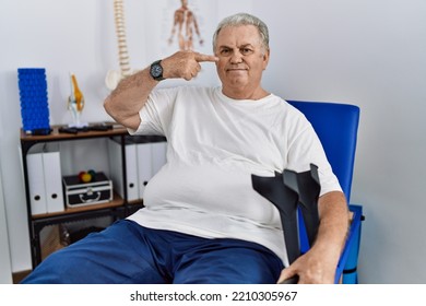 Senior Caucasian Man At Physiotherapy Clinic Holding Crutches Pointing With Hand Finger To Face And Nose, Smiling Cheerful. Beauty Concept 