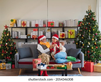 Senior Caucasian Man In Santa Claus Hat Giving Christmas Gift To His Wide Couple While Sitting On Sofa Couch With Decorated Christmas Tree In Living Room At Home. Happy Surprise Gift From Husband. 