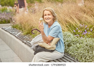 Senior Caucasian Pretty Lady About 60 Years Old Is Sitting On The Bench In The Public Park And Eating Ice Cream. She Is Smiling And Looks Cheerful And Happy. There Are Flowers On The Background.