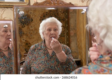 Senior Caucasian Woman About Ninety Years Old Puts On Lipstick Before A Three Sided Mirror In Her Bed Room