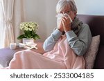 Senior caucasian woman blowing nose with severe cold and fever symptoms like seasonal flu, covid or pollen allergy - woman self-medicate on couch at home with pills, thermometer and blanket