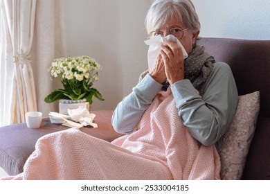 Senior caucasian woman blowing nose with severe cold and fever symptoms like seasonal flu, covid or pollen allergy - woman self-medicate on couch at home with pills, thermometer and blanket - Powered by Shutterstock