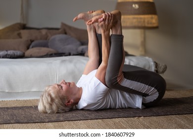 Senior Caucasian Woman Doing Yoga In Sunny Room. Happy Baby Pose.