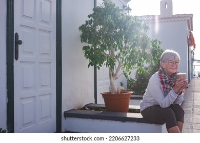 Senior caucasian woman holding coffee cup sitting outdoor home, looking away. Elderly female having calm peaceful morning relaxing outside with mug, drinking coffee - Powered by Shutterstock