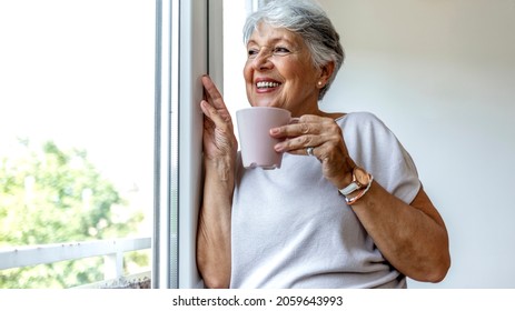 A senior Caucasian woman leans against a window frame while looking outside thoughtfully. She is holding a cup of tea in her hand while smiling in casual clothes. Old woman looking through the window. - Powered by Shutterstock