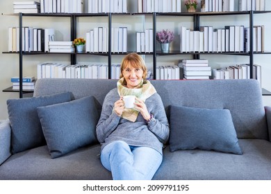 Senior Caucasian Woman Sitting Relaxingly On The Sofa Couch Watching Movie During Winter Time With Warm Cloth And Scarf In Her Cozy Home Living Room