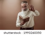 Senior charismatic man with a gray beard in a light shirt holding Holy Bible book and preaching