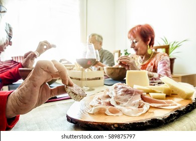 Senior Cheerful Friends Eating Appetizer Lunch With Cheese,ham And Salami At Home - Old People Having Fun Toasting Wine  - Joyful Elderly Active Concept - Focus On Left Bottom Hand - Warm Filter