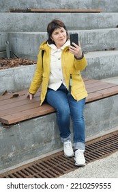 Senior Citizen Brunette Woman Sits In City Park On The Steps, Smiling While Talking On Video Call. Woman Holds Smartphone In Front Of Her. Woman In A Yellow Jacket Sits On A Bench To Talk On The Phone