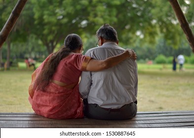 Senior Citizen Indian Woman Reads From Mobile And With Her Hand Over Her Husband's Shoulder Shot From Behind While Sitting In A Park In New Delhi, India. Concept Love