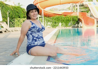 Senior Citizen In Swimsuit Sits On The Side Of The Pool And Put Her Feet In The Water. Mature Woman In Panama Is Relaxing In Hotel. Woman Smiles And Looks At The Camera.