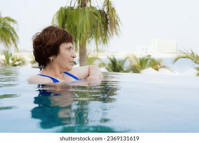 Senior citizen woman relaxes in the infinity pool. Portrait of a retired woman on vacation in swimsuit. Woman looking at the sea from an infinity pool - Powered by Shutterstock