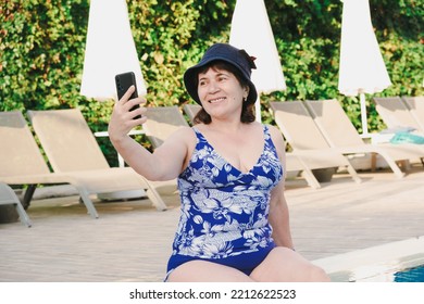 Senior Citizen Woman In Swimsuit Sits On The Side Of The Pool, Put Her Feet In The Water And Takes A Selfie On A Smartphone. Mature Woman Is Relaxing In Hotel. Woman Smiles