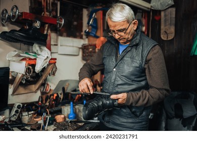 A senior cobbler is standing at cobbler workshop, using scissors and repairing boots. Portrait of a focused senior shoemaker repairing boots at his small workshop. Small business owner and craftsman. - Powered by Shutterstock