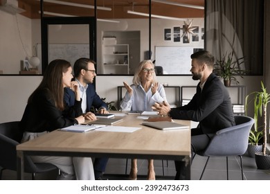 Senior company owner woman talking to team of younger managers on corporate meeting, sitting at boss place at large table, explaining teamwork strategy, smiling, discussing collaboration