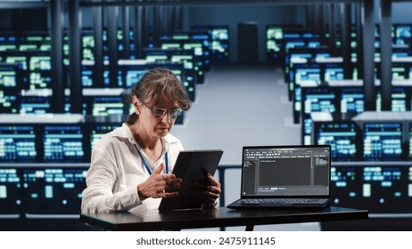 Senior computer scientist in server farm completing tasks on tablet while running scripts on laptop terminal. Expert amidst supercomputers manipulating code and improving cybersecurity