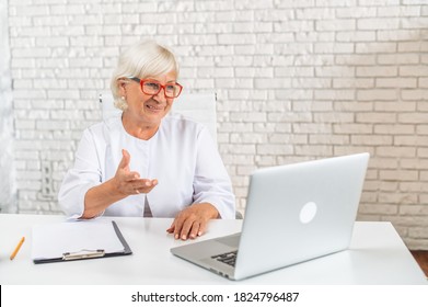 Senior Confident Female Doctor In Eyeglasses Sits In The Cab And Using Laptop For Online Consultation. A Gray Haired Woman In White Formal Coat Looks At Webcam And Talking. Virtual Visit Concept