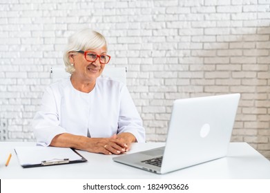 Senior Confident Female Doctor In Eyeglasses Sits In The Cab And Using Laptop For Online Consultation. A Gray Haired Woman In White Formal Coat Looks At Webcam And Talking. Virtual Visit Concept