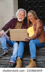 Senior Couple Of Asian Man And African American Woman Using Computer Outdoors At Dusk. - Retirement Concept -