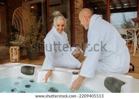 Senior couple in bathrobe checking temperature in outdoor hot tub, preparing for bathing with his wife.