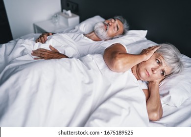 Senior couple in the bed. Pretty aged woman is angy with her sleeping husband covering ears with hands. - Powered by Shutterstock