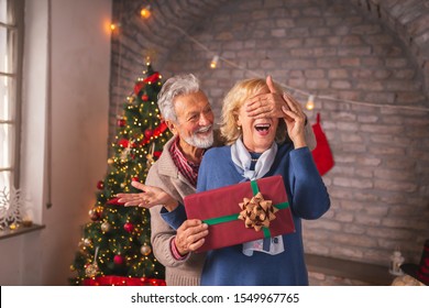 Senior couple celebrating Christmas at home, exchanging Christmas presents; husband holding his wife's eyes closed, while surprising her with a Christmas gift - Powered by Shutterstock