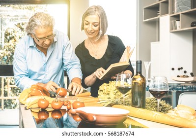 Senior Couple Cooking Healthy Food And Drinking Red Wine At House Kitchen - Retired People At Home Preparing Lunch With Recipe Book About Bio Vegetables - Happy Elderly Concept With Mature Pensioner 