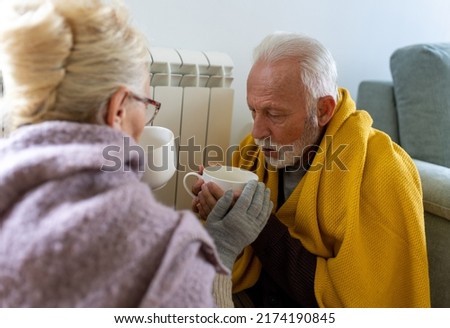 Senior couple covered with blanket sitting beside heater in living room, drinking hot tea from big cup and trying to warm in winter season and energy crisis