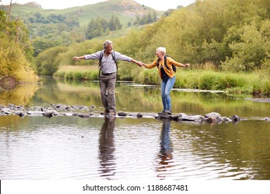 Senior Couple Crossing River Whilst Hiking In UK Lake District