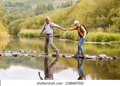 Senior Couple Crossing River Whilst Hiking In UK Lake District