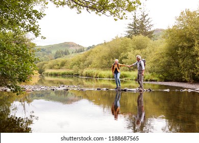 Senior Couple Crossing River Whilst Hiking In UK Lake District