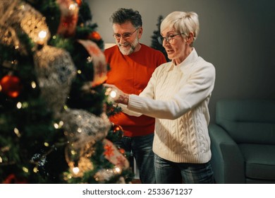 Senior couple is decorating the christmas tree at home. They are both wearing glasses and sweaters and are smiling as they put the finishing touches on the tree - Powered by Shutterstock