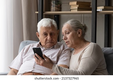 Senior Couple Discuss Mobile App Seated On Sofa At Home, Staring At Smart Phone Screen Learn New Software. Loving Wife Explains Modern Device Usage To Mature Husband. Help, Tech And Older Gen Concept