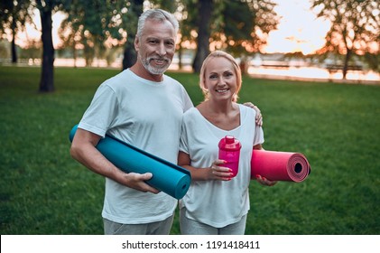 Senior couple is doing sport outdoors. Standing with yoga carpet and shaker in hands in park during sunrise. - Powered by Shutterstock