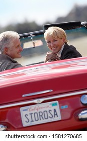 Senior Couple Driving Classic Red Convertible Car On Road Trip At Camera