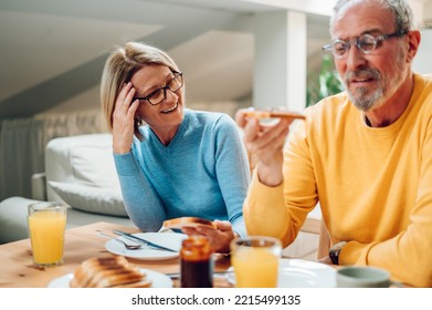 Senior Couple Eating Breakfast At Home And Sitting At The Table. Focus On A Mature Woman Laughing To Her Husband Joke. Happy Aged Husband And Wife Enjoy Spending Time At Home Together. Copy Space.