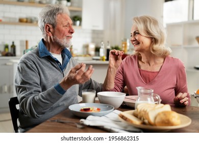 Senior Couple Eating Breakfast In The Kitchen. Husband And Wife Talking And Laughing While Eating A Sandwich	