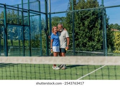 senior couple embraces on the padel court, looking at the camera with joyful expressions while holding their paddles - Powered by Shutterstock