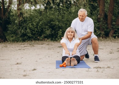 Senior couple engaging in yoga exercise outdoors, showcasing fitness, health, and togetherness in a natural setting. A man helps a woman do exercises. - Powered by Shutterstock